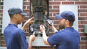 Two men electricians working on a residential breaker box.  They use tools to assess the repair and wear matching blue uniforms.  The multi-ethnic group is discussing next steps in the job repair.  Electric meter to side.